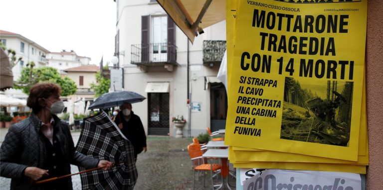 Menschen stehen vor einem Zeitungskiosk, an dem eine Zeitung mit der Schlagzeile „Mottarone-Tragödie mit 14 Toten“ ausgehängt ist. Beim Absturz einer Gondel einer Seilbahn am norditalienischen Lago Maggiore am 23.05.2021 haben mindestens 14 Menschen ihr Leben verloren.