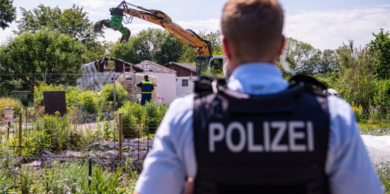 Ein Polizist beobachtet den Abriss der Gartenlaube des Verdächtigen im Missbrauchsfall von Münster mit einem Bagger. In der Laube des Kleingartenvereins sollen mehrere Männer zwei Jungen schwer missbraucht haben.