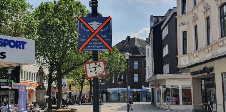 Ein blaues Schild am Rande der Fußgängerzone, auf dem Maskenpflicht steht, ist mit einem großen X aus rotem Klebeband quasi durchgestrichen.
