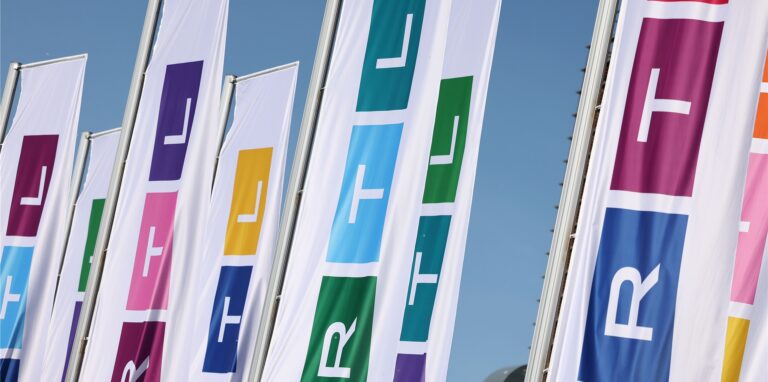 Fahnen mit dem neuen Logo des Fernsehsenders RTL wehen vor dem Gebäude. Die drei Buchstaben des Privatsenders sind schmaler, statt aus den drei Grundfarben Rot, Gelb, Blau sind die drei Blöcke des Logos aus allen möglichen Farben zusammensetzen.