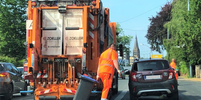 Ein Müllwerker zieht eine blaue Tonne hinter sich her, hinter ihm steht ein Müllfahrzeug.