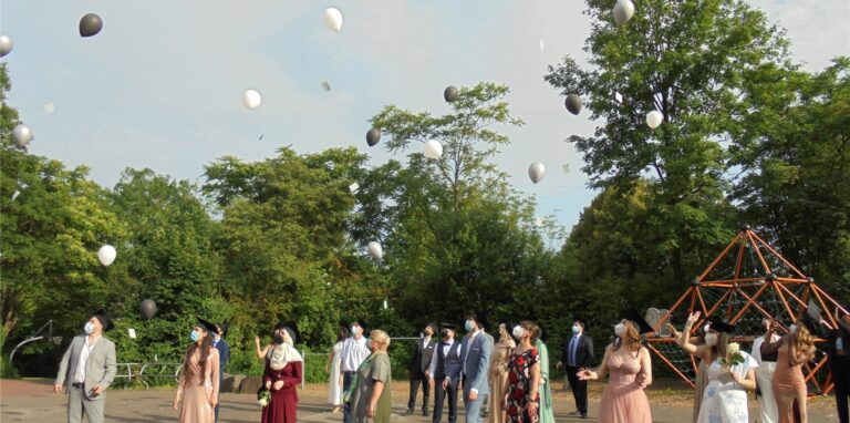 Die Abiturienten und Abiturientinnen der Rosa-Parks-Schule, die alle einen schwarzen Absolventenhut mit Quaste tragen, lassen zur Feier des Tages Ballons aufsteigen, an denen Karten mit guten Wünschen befestigt sind.