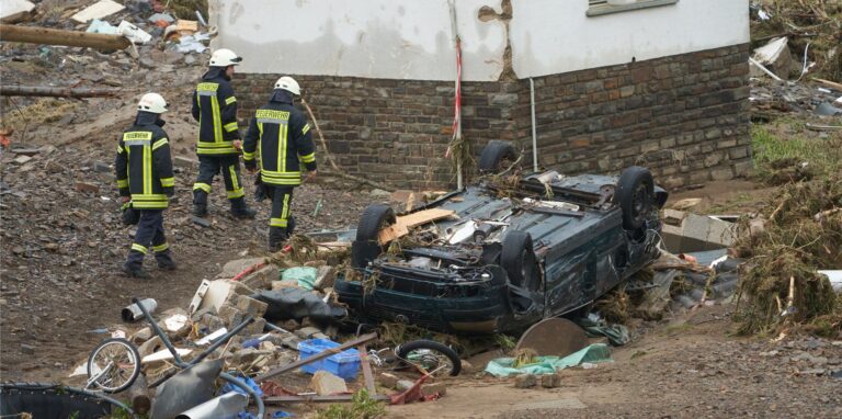Feuerwehrleute gehen in der Gemeinde Schuld an einem zerstörten Auto vorbei.