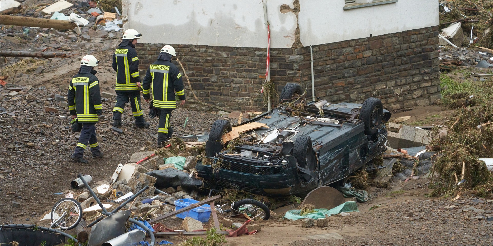 Feuerwehrleute gehen in der Gemeinde Schuld an einem zerstörten Auto vorbei.