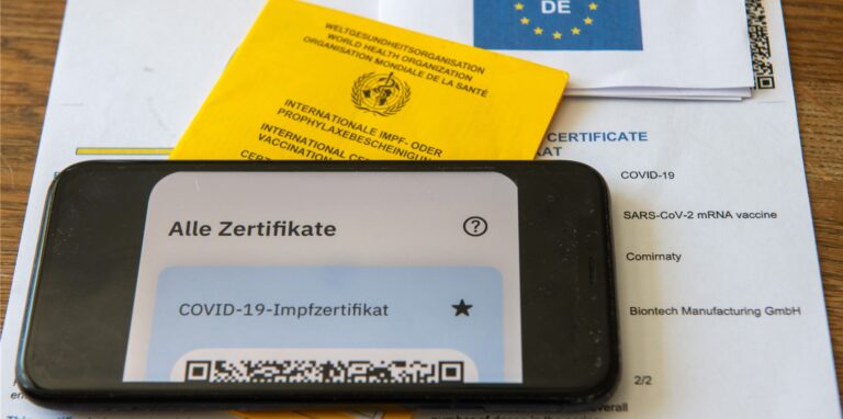 Ein Impfpass und ein Smartphone, auf dem die App CovPass läuft, liegen auf einem Impfzertifikat, das von einer Apotheke ausgestellt wurde.