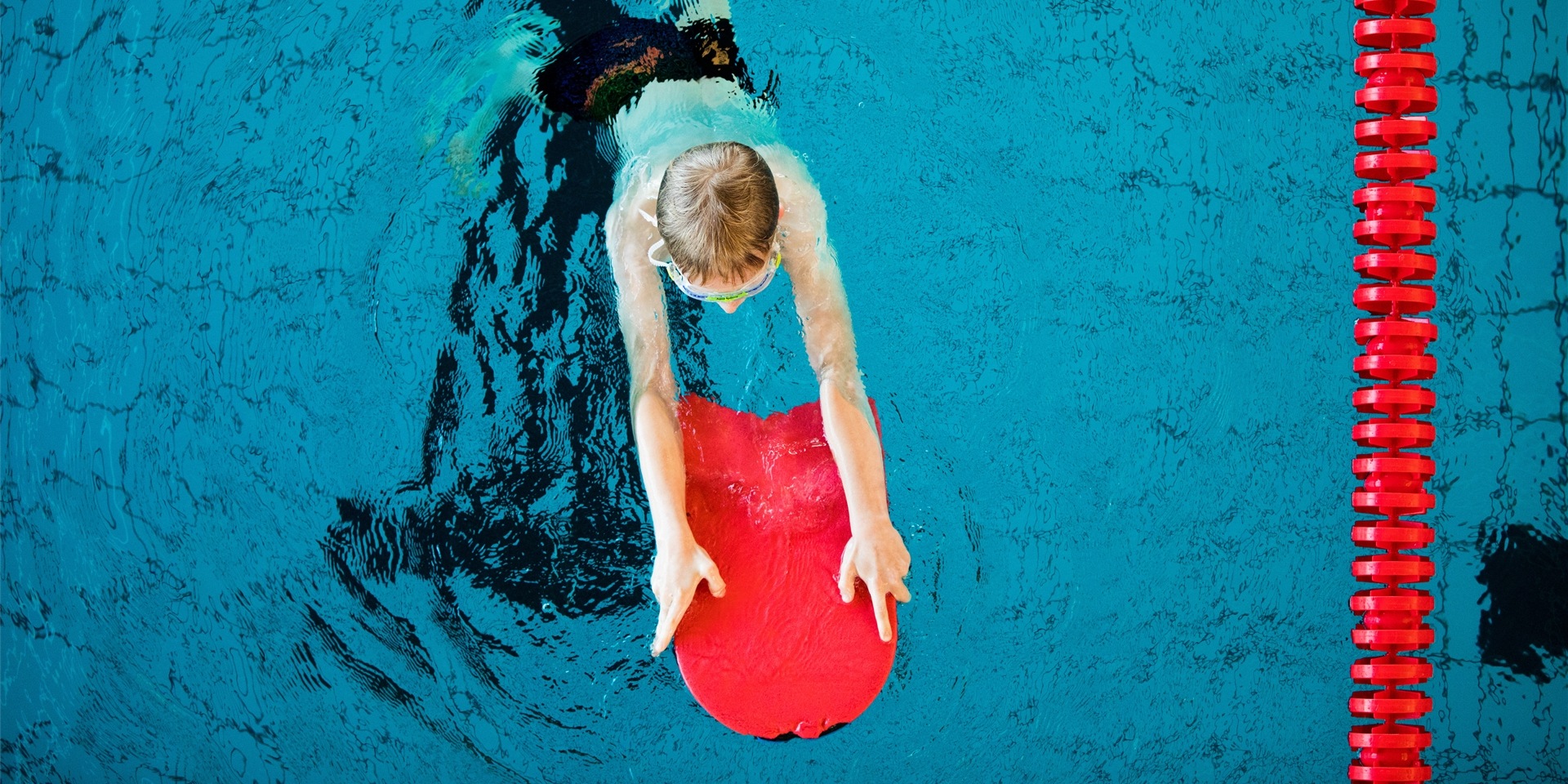 Blick von oben auf einen schwimmenden Jungen, der ein Schwimmbrett in den Händen hält. (Symbolbild)