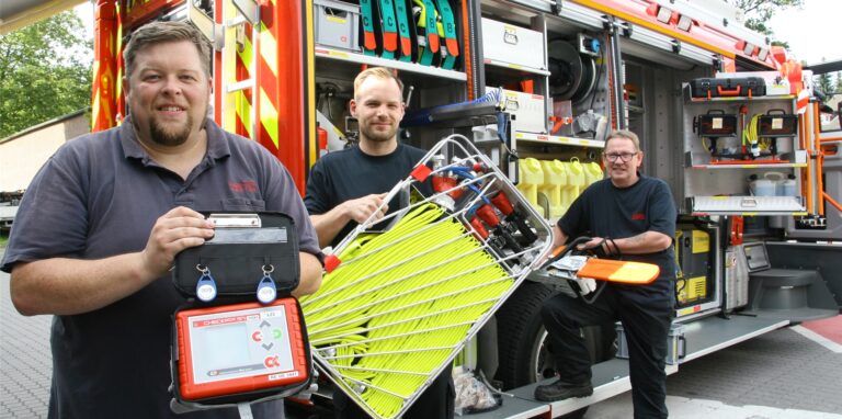 Vor dem neuen Löschfahrzeug zeigen (v.l.) Dennis Piechkamp, Florian Piechkamp und Uli Würfel drei wichtige Ausrüstungsgegenstände: ein neues Gerät zur digitalen Überwachung der Einsatzkräfte, die Atemschutzgeräte tragen; einen Tragekorb mit leichten, dünnen D-Schläuchen, die bei Waldbränden hilfreich sind; eine Motorsäge, von der es angesichts der immer häufigeren Sturm-Einsätze jetzt zwei auf dem Fahrzeug gibt.