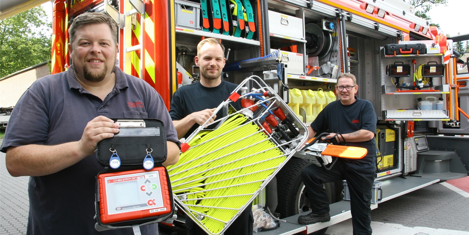 Vor dem neuen Löschfahrzeug zeigen (v.l.) Dennis Piechkamp, Florian Piechkamp und Uli Würfel drei wichtige Ausrüstungsgegenstände: ein neues Gerät zur digitalen Überwachung der Einsatzkräfte, die Atemschutzgeräte tragen; einen Tragekorb mit leichten, dünnen D-Schläuchen, die bei Waldbränden hilfreich sind; eine Motorsäge, von der es angesichts der immer häufigeren Sturm-Einsätze jetzt zwei auf dem Fahrzeug gibt.