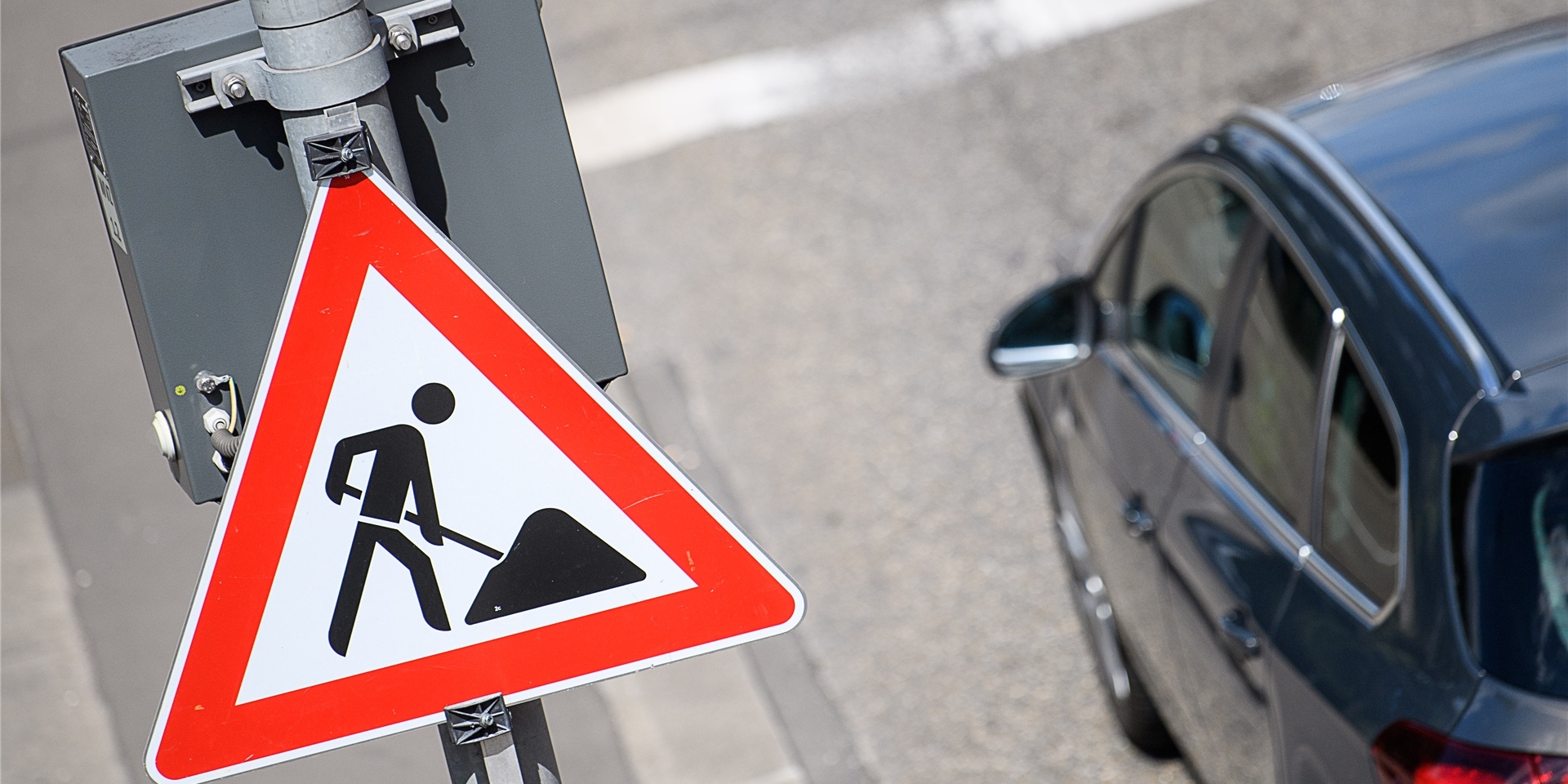Das Foto zeigt ein Baustellenschild, im Hintergrund ist in der rechten Bildhälfte ein schwarzes Auto auf dem Asphalt erkennbar.