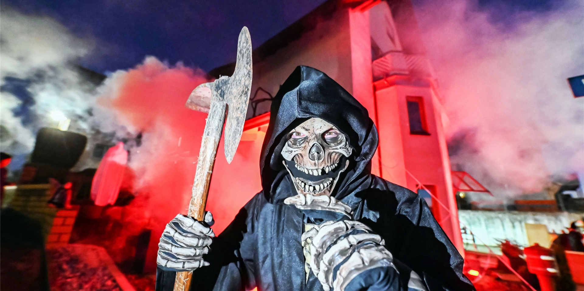 Ein Mann im schwarzen Umhang und Totenschädel sowie Skeletthänden steht mit einer Axt vor einem rot erleuchteten Haus.