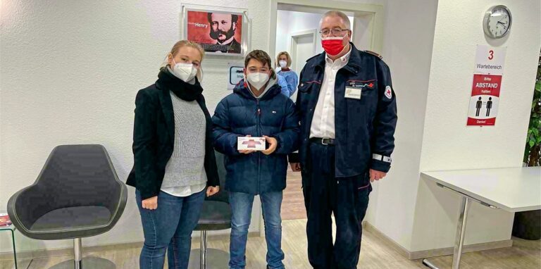 Juliana Gabriel (Stadt Herten) und Ralph Hoffert (DRK Herten) begrüßen den 14-jährigen Filippo Calamini im Impfzentrum. Als Erster, der sich am Freitag seine Erstimpfung abholt, erhält er eine Tafel Schokolade als Dankeschön.