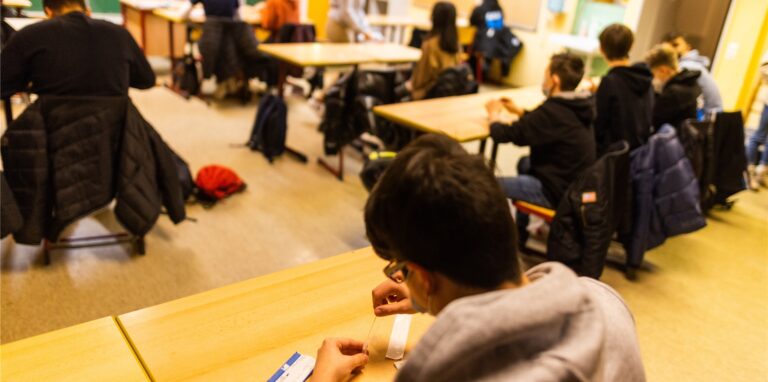Ein Schüler einer Realschule hält sein Probestäbchen in die Testflüssigkeit für einen Corona-Schnelltest, während im Hintergrund andere Schülerinnen und Schüler zu sehen sind. Hunderte Schüler in NRW lehnen die Tests aber ab und verweigern somit den Präsenzunterricht.