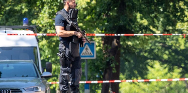 Ein Polizist mit Schutzweste und Gewehr steht an einer Absperrung in Espelkamp. In Espelkamp sind nach Angaben der Bielefelder Polizei zwei Menschen erschossen worden.