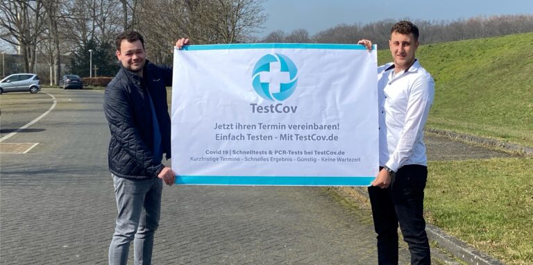 Zwei Mitarbeiter der Drive in Corona-Teststelle der Schreurs Consulting Group halten auf dem Atlantis-Parkplatz am Lippedeich ein Plakat mit Informationen über das Testzentrum hoch.