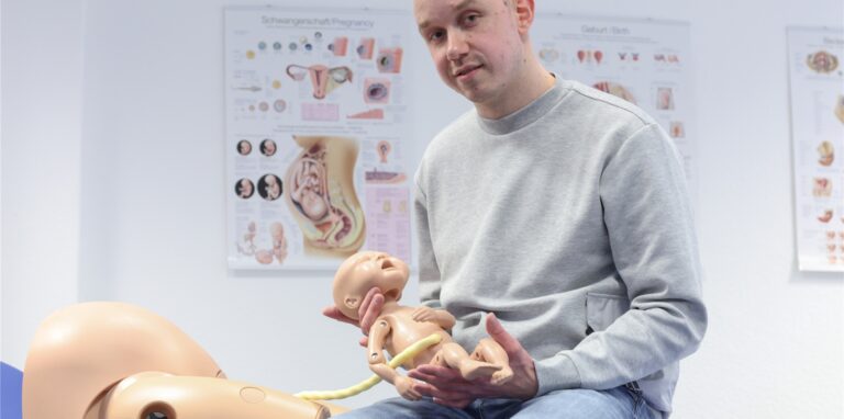 Jonas Küppers, männliche Hebamme, hält eine Neugeborenen-Übungspuppe mit Nabelschnur. Küppers absolviert ein duales Studium, um den Beruf der Hebamme zu lernen.