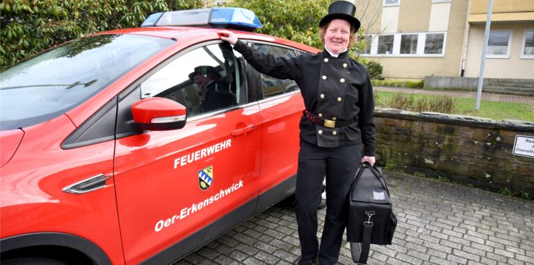 Schornsteinfegerin Sandra Zollhofer steht in Arbeitskleidung neben einem Dienstfahrzeug der Feuerwehr.