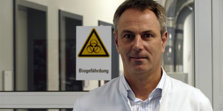 Dr. Frank Hünger im weißen Kittel in einem Arbeitsbereich. Im Hintergrund ist ein Schild mit der Aufschrift „Biogefährdung“ zu sehen.