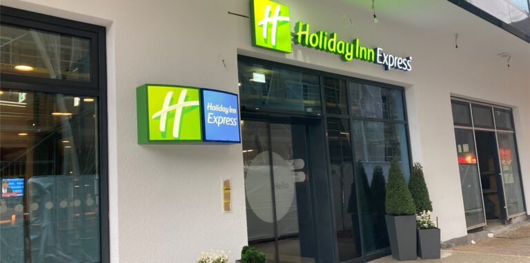 Eingangsbereich des Hotels Holiday Inn Express im Marktquartier an der Herrenstraße in der Innenstadt von Recklinghausen. Links und rechts neben dem gläsernen Eingangsbereich stehen Topfpflanzen, darüber hängt die grüne Beleuchtung mit dem Namenszug des Hotels.