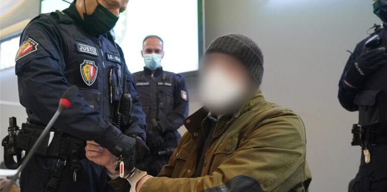 Ein Justizbeamter entfernt die Handschellen des Angeklagten (2.v.r) im Sitzungssaal der Außenstelle des Landgerichts zu Beginn des Prozesstages wegen eines Dreifachmordes.