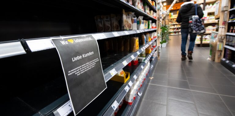 Eine Frau läuft durch einen Supermarkt. Im Vordergrund ist ein leeres Regal zu sehen, in dem normalerweise Mehl steht.