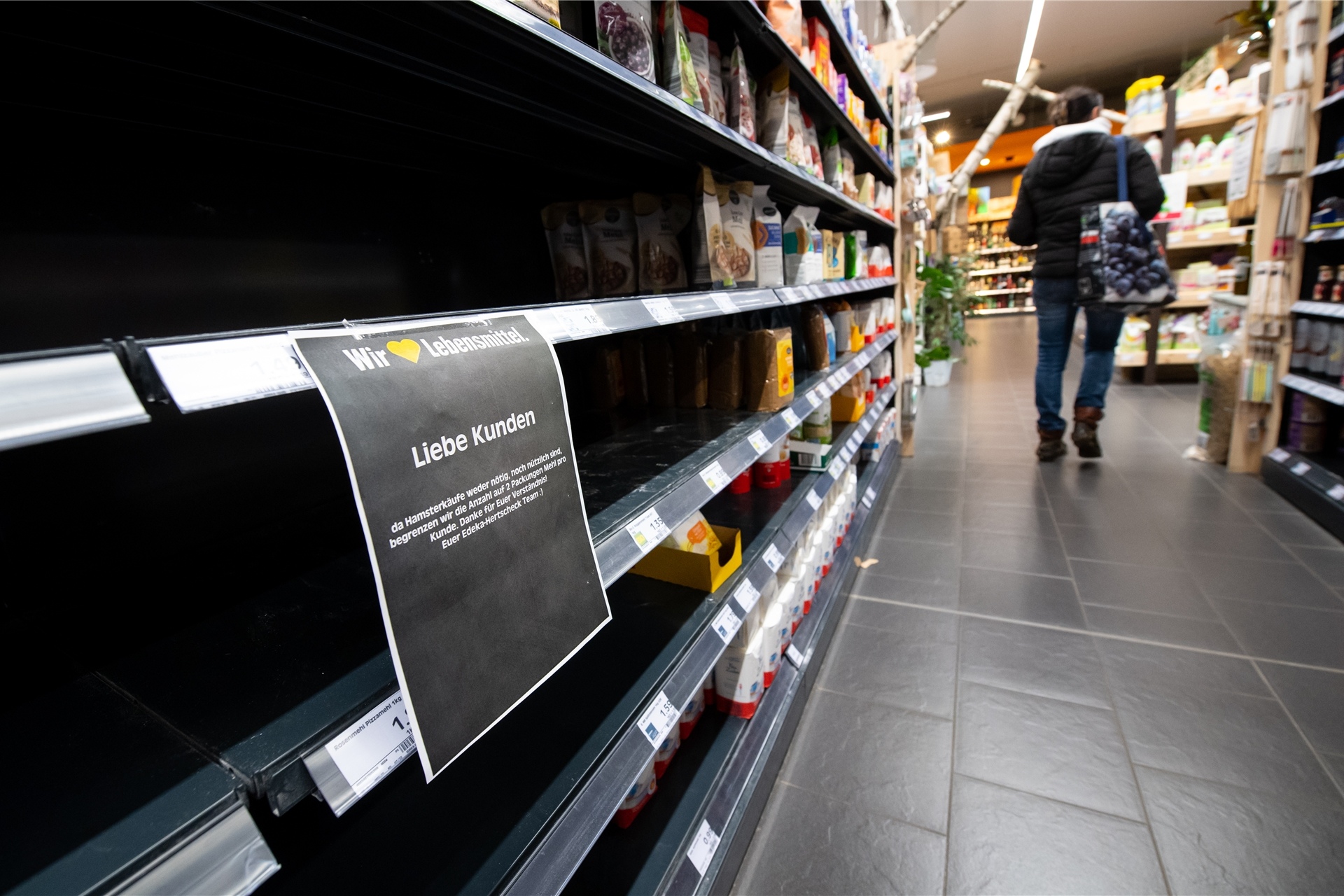 Eine Frau läuft durch einen Supermarkt. Im Vordergrund ist ein leeres Regal zu sehen, in dem normalerweise Mehl steht.