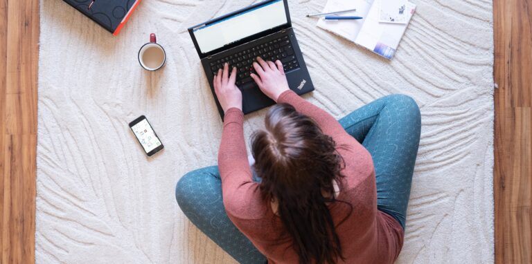 Eine Frau sitzt im Homeoffice auf einem Teppich und arbeitet. Neben dem Laptop steht ein Kaffee.