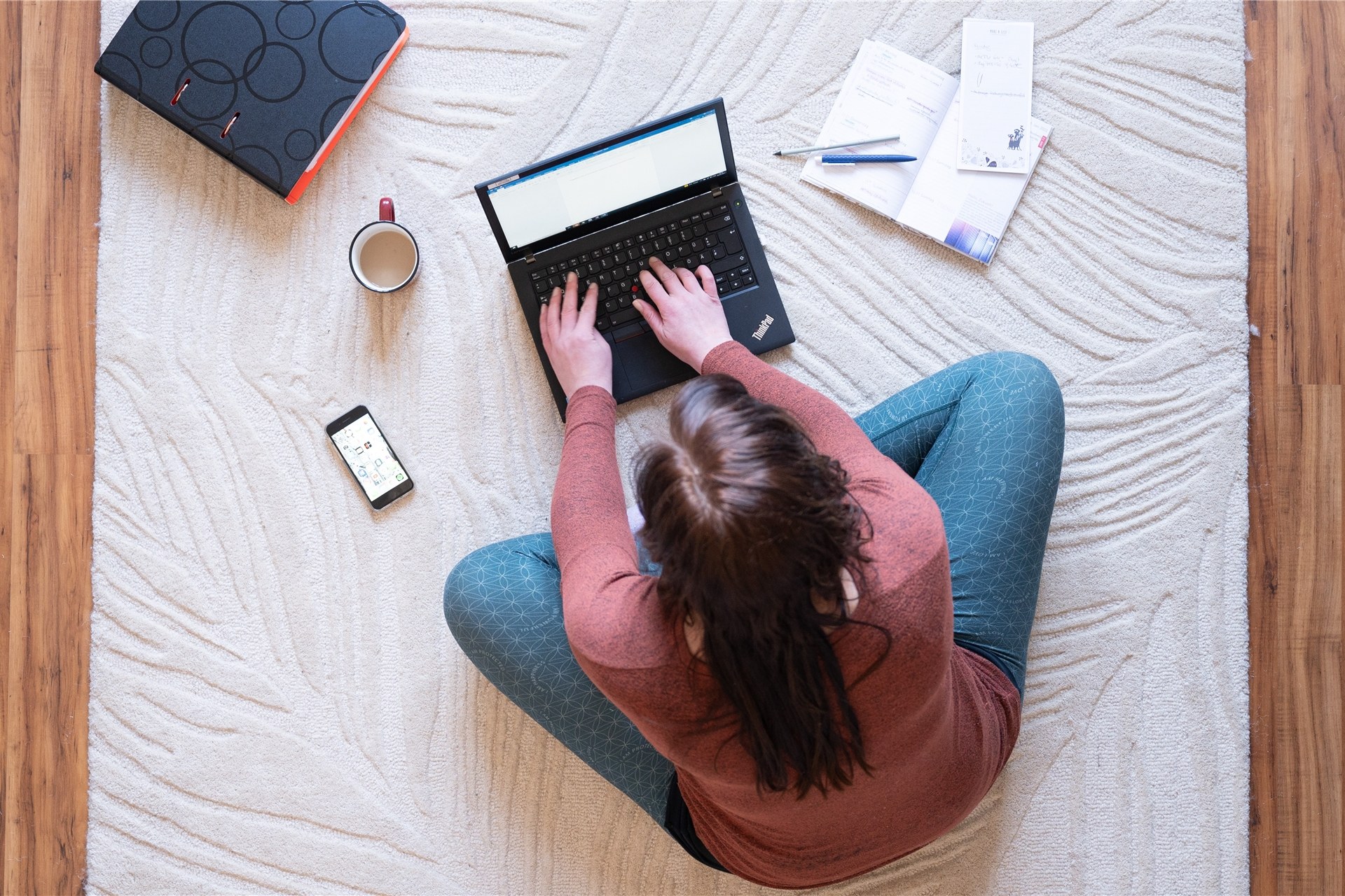 Eine Frau sitzt im Homeoffice auf einem Teppich und arbeitet. Neben dem Laptop steht ein Kaffee.