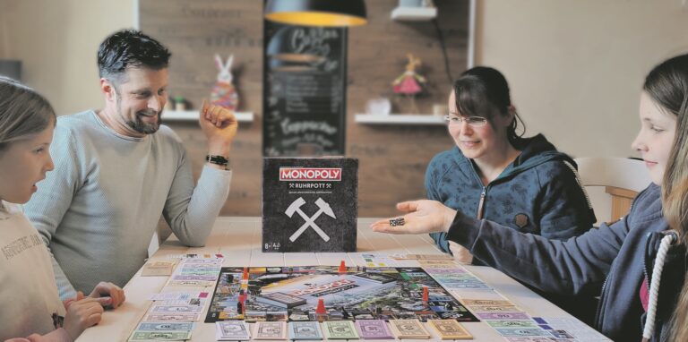 Vier Personen – vermutlich Vater, Mutter und zwei Töchter – spielen an einem Tisch sitzend das Brettspiel Ruhrpott-Monopoly. Das Mädchen rechts ist im Begriff, die Würfel in ihrer Hand zu werfen.