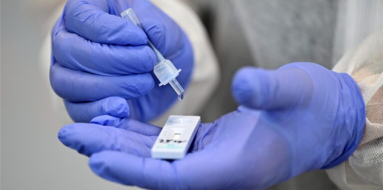 Zwei Hände, die in blauen Schutzhandschuhen stecken, halten einen Coronatest.