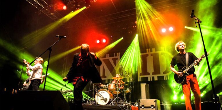 Drei Mitglieder der Waltroper Band „No ID“ stehen mit ihren Instrumenten im Scheinwerferlicht auf einer Bühne, im Hintergrund das Band-Logo.
