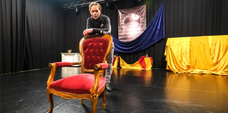 Ein Mann steht hinter einem roten Stuhl auf der Theaterbühne.