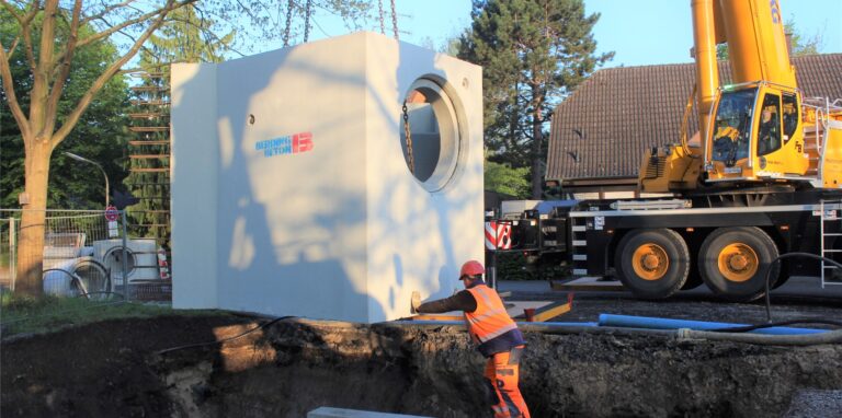Ein Kran hievt ein Betonbauteil für die Kanalisation in eine Baugrube, ein Bauarbeiter stützt das Teil.