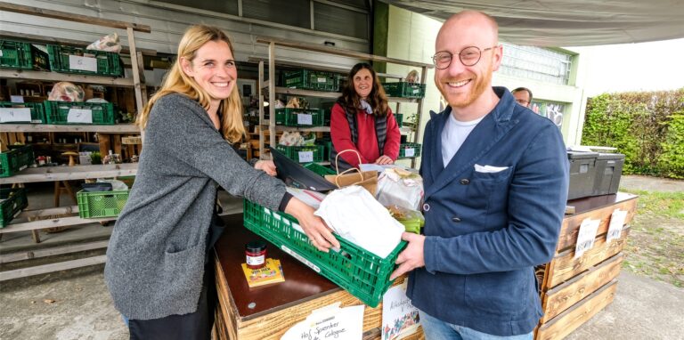 Sarah Küper von den Marktschwärmern Recklinghausen-Nord steht links im Bild und reicht ihrem Kunden Markus Großbröhmer seine mit Lebensmitteln gefüllte grüne Plastikkiste über die Verkaufstheke. Im Hintergrund steht zwischen den beiden Andrea Kampmann vom Hof Kampmann aus Dortmund-Mengede.