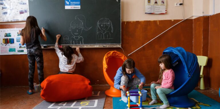 Kinder spielen in einem Klassenraum auf dem Boden, andere malen auf der Tafel mit Kreide.