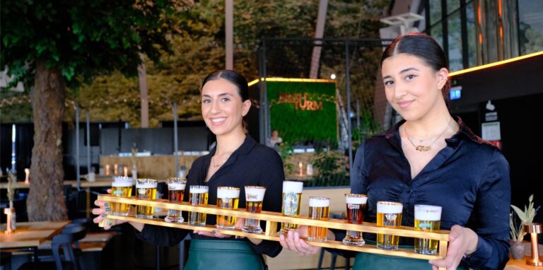 Zwei Mitarbeiterinnen des „Brauturm“ halten ein Holzgestell mit elf Biergläsern in den Händen.