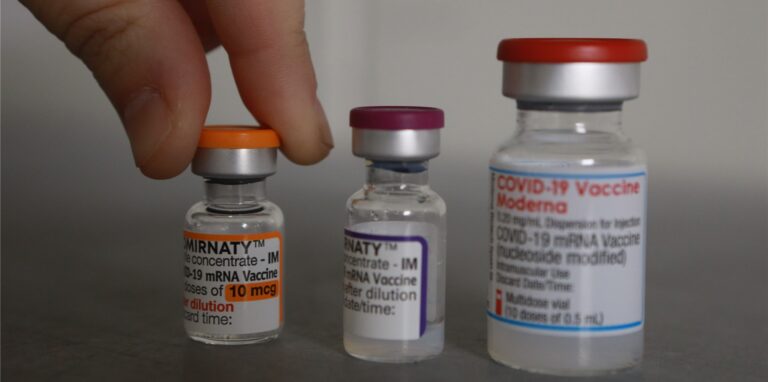 Drei Flaschen mit den Impfstoffen gegen Covid-19, Comirnaty, Moderna und Biontech/Pfizer, stehen nebeneinander; eine Hand hebt die linke Ampulle an.