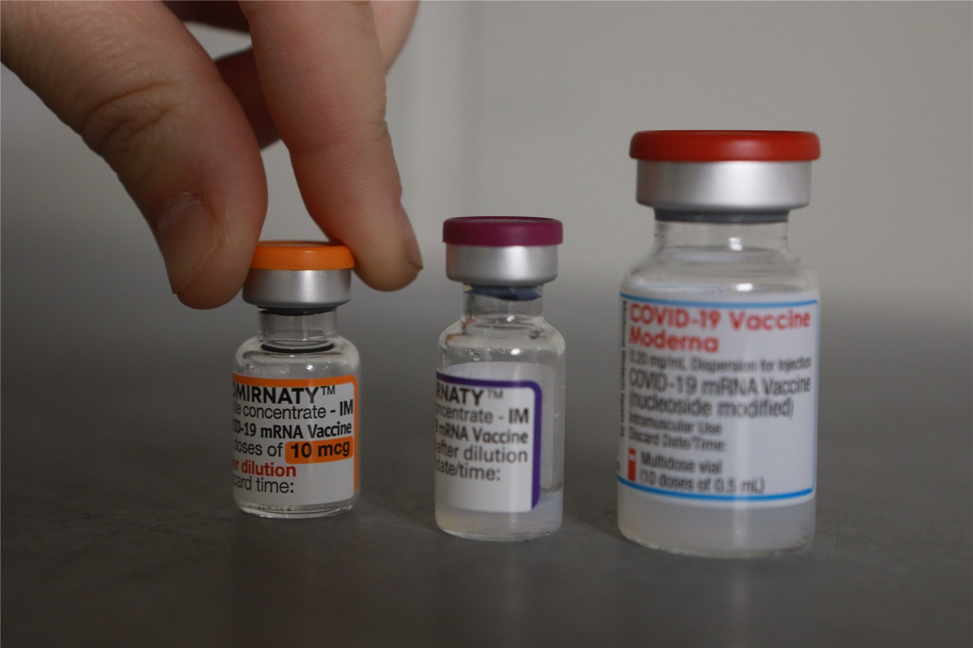Drei Flaschen mit den Impfstoffen gegen Covid-19, Comirnaty, Moderna und Biontech/Pfizer, stehen nebeneinander; eine Hand hebt die linke Ampulle an.