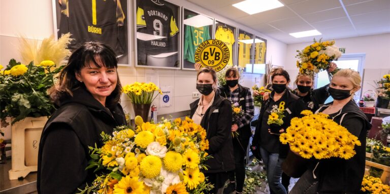 Birgit Honvehlmann (l.) und fünf Kolleginnen stehen mit verschiedenen Sträußen in der Hand vor einer Reihe von gerahmten BVB-Trikots.