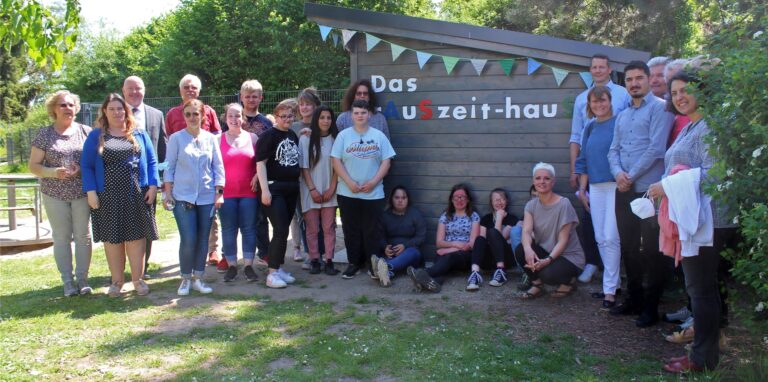 Eine Gruppe von Menschen steht vor einem Gartenhütten-ähnlichen Haus, auf dem „Rauszeithaus“ steht.