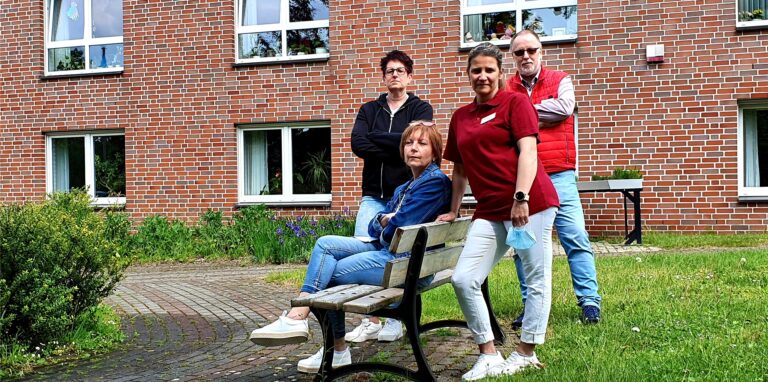 Drei Frauen und ein Mann sitzen in einem Park auf einer Bank oder stehen daneben.