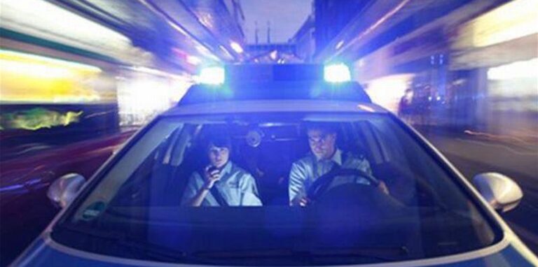 Zwei Polizisten sitzen in einem Streifenwagen mit angeschaltetem Blaulicht.