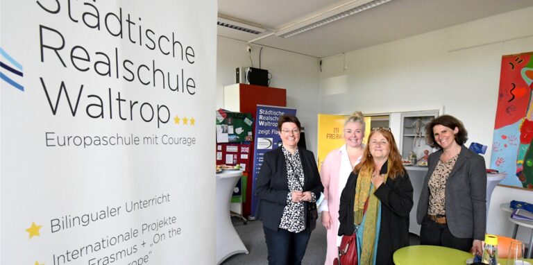 Vor einem Plakat mit der Aufschrift „Städtische Realschule Waltrop – Europaschule mit Courage“ stehen Schulleiterin Lisa Dahlhues, ihre Stellvertreterin Hania Kampa-Jansing sowie Sabine Volkner und Kirsten Laroussi von der Europakommission aus dem Bildungs-Ministerium.