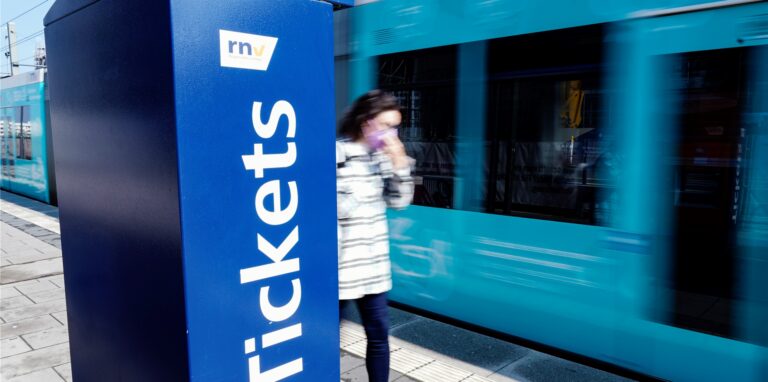 Eine Straßenbahn fährt an einer Straßenbahnhaltestelle an einem Ticketautomaten vorbei.