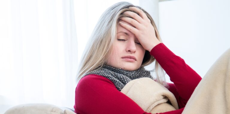 Eine Frau liegt unter einer Decke eingekuschelt auf der Couch, hat die Augen zu und hält sich die Hand an den Kopf.