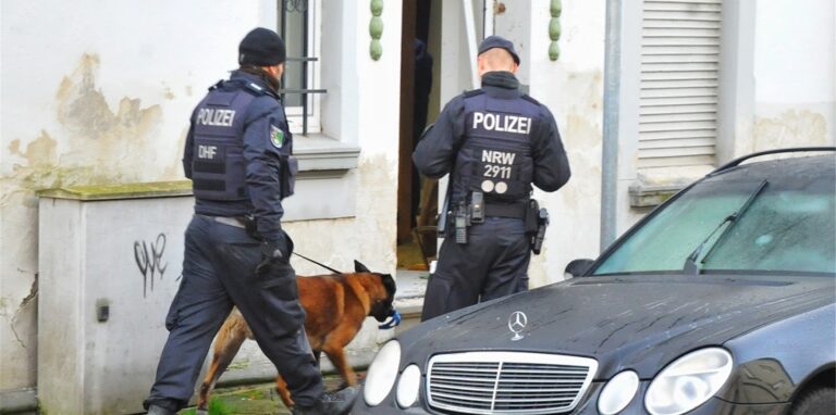 Polizisten und ein Spürhund stehen vor einem Haus.
