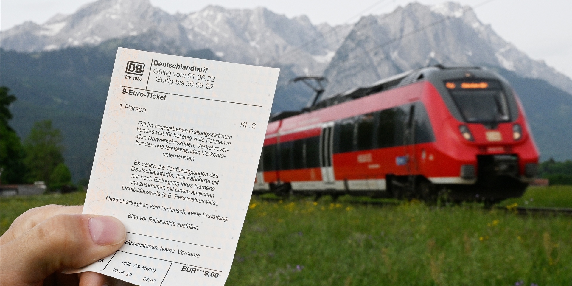 Ein Mann hält ein 9-Euro-Ticket in der Hand, im Hintergrund fährt ein Zug der Deutschen Bahn.