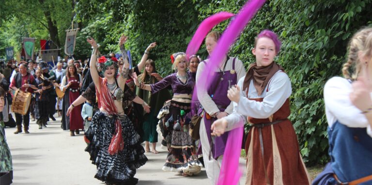 Mittelalterlich gekleidete Personen, unter anderem Bauchtänzerinnen und Frauen, die Bänder schwenken, laufen durch den Moselbachpark Waltrop.