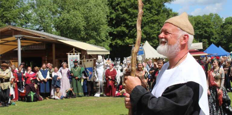 Fest-Organisator Detlef Huß steht, gestützt auf einen Stab, in Mittelalter-Kleidung auf einer Wiese, im Hintergrund weitere Mitwirkende des Mittelalterlich Gaudium in Waltrop.