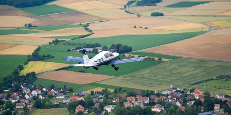 Ein Kleinflugzeug fliegt über eine Siedlung und Felder.