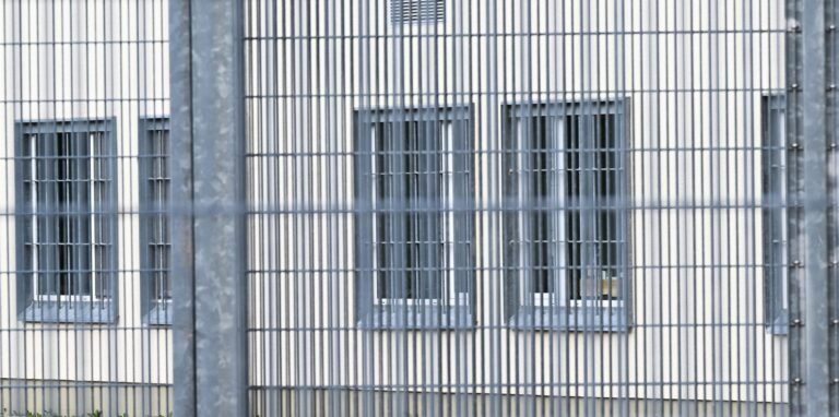 Blick durch einen Zaun auf vergitterte Fenster
